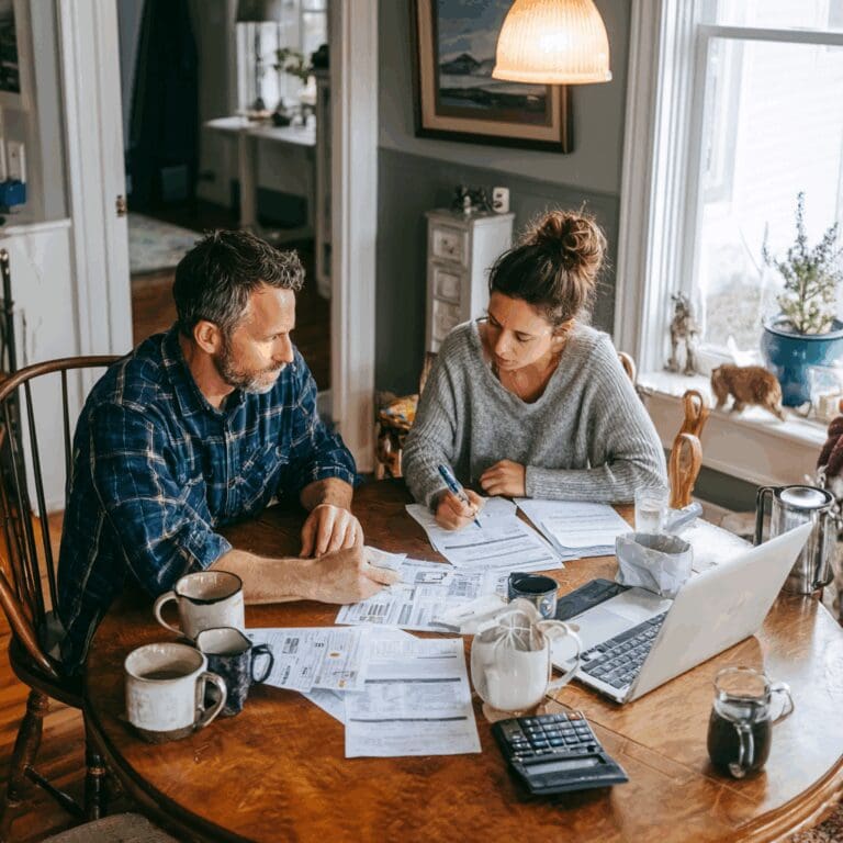 Self-employed couple sitting at their kitchen table in a BC home, reviewing mortgage documents and financial paperwork together with a laptop, calculator, and coffee mugs in view.